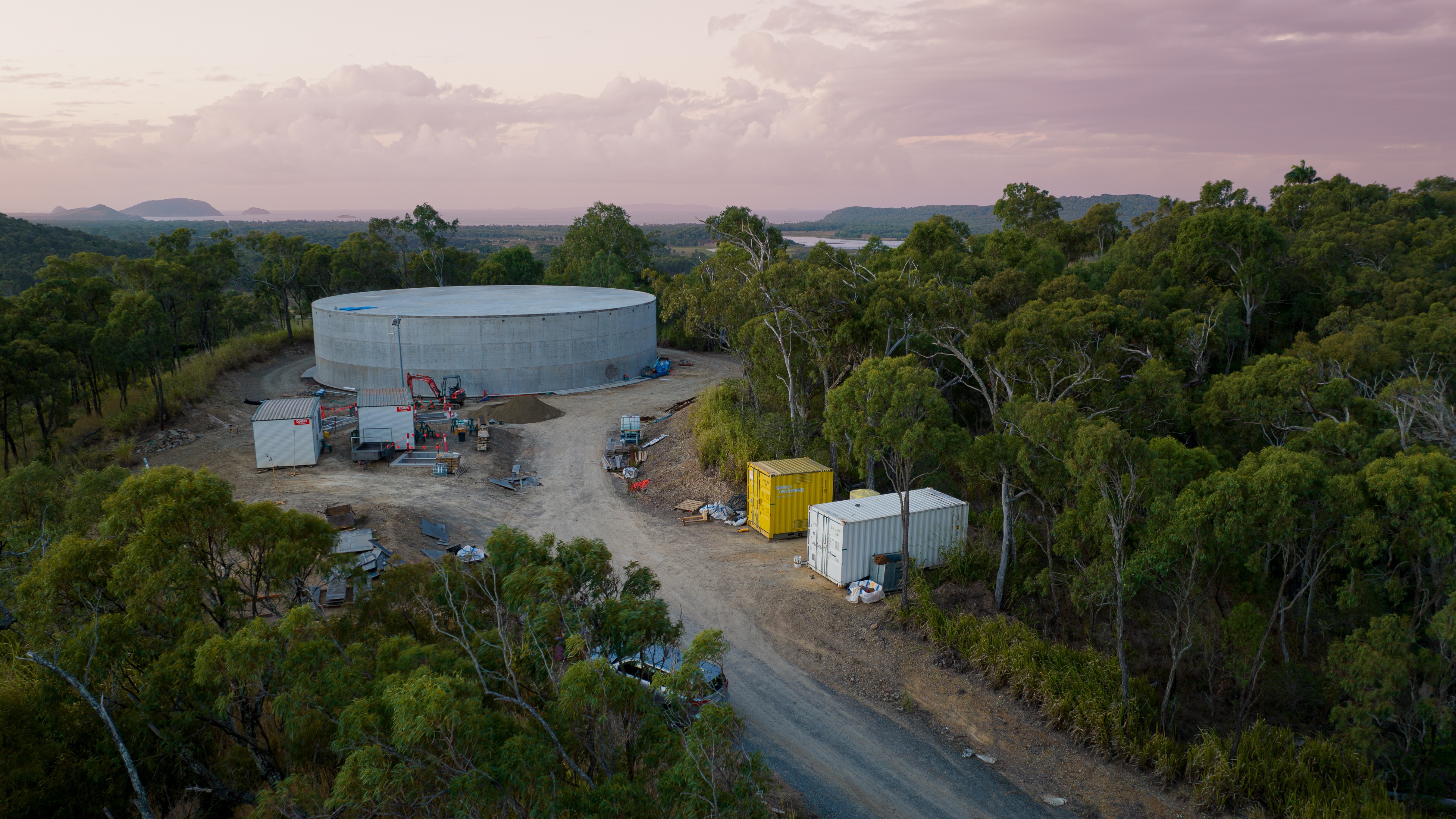 Emu Park Reservoir construction site in a forested area with a large cylindrical concrete structure, surrounded by equipment and shipping containers at sunrise or sunset.