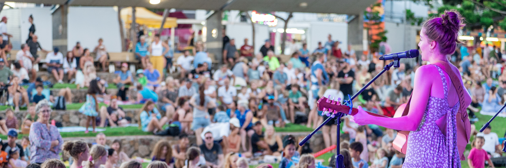 Outdoor crowd watching a guitarist perform at a festival, with tents and grass terraces in view.