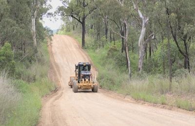 Yellow grader driving on a dirt road through a forested area.