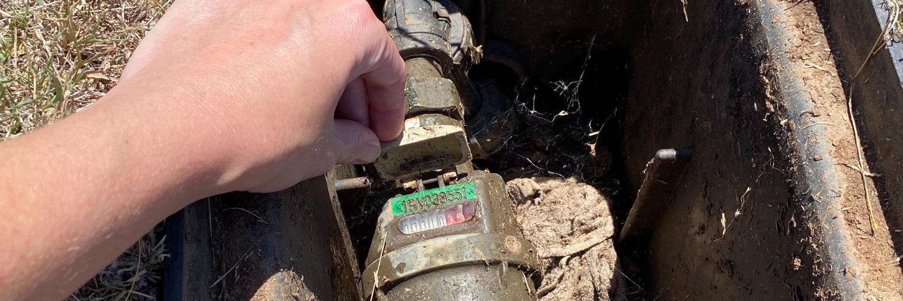 Person's hand adjusting a water meter outdoors, partially buried in soil and surrounded by grass.