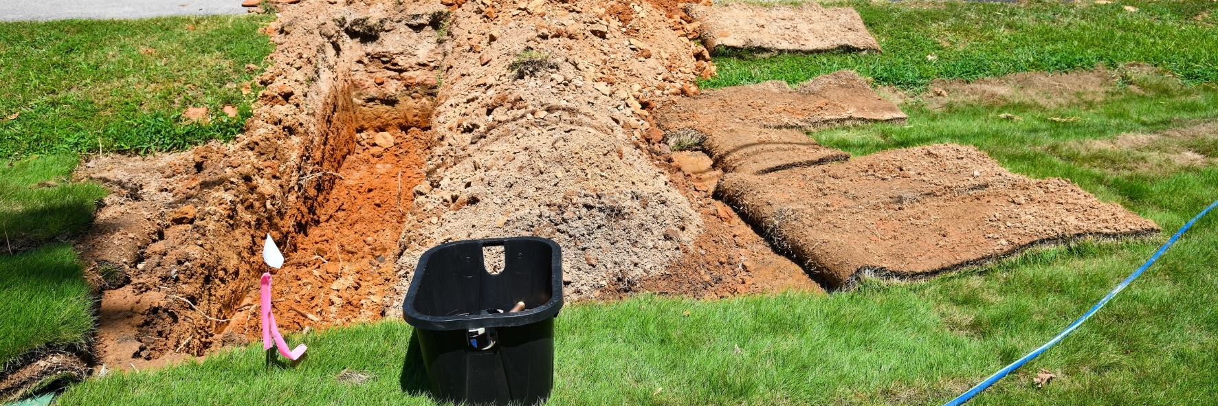 Construction site with a trench and traffic cones, showing a black container and pink pipe near the excavation.