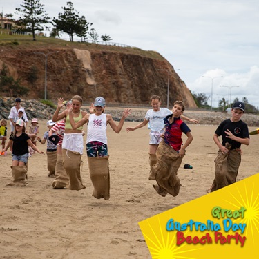 Children in a sack race on a beach during an Australia Day event, with cliffs and a road in the background.