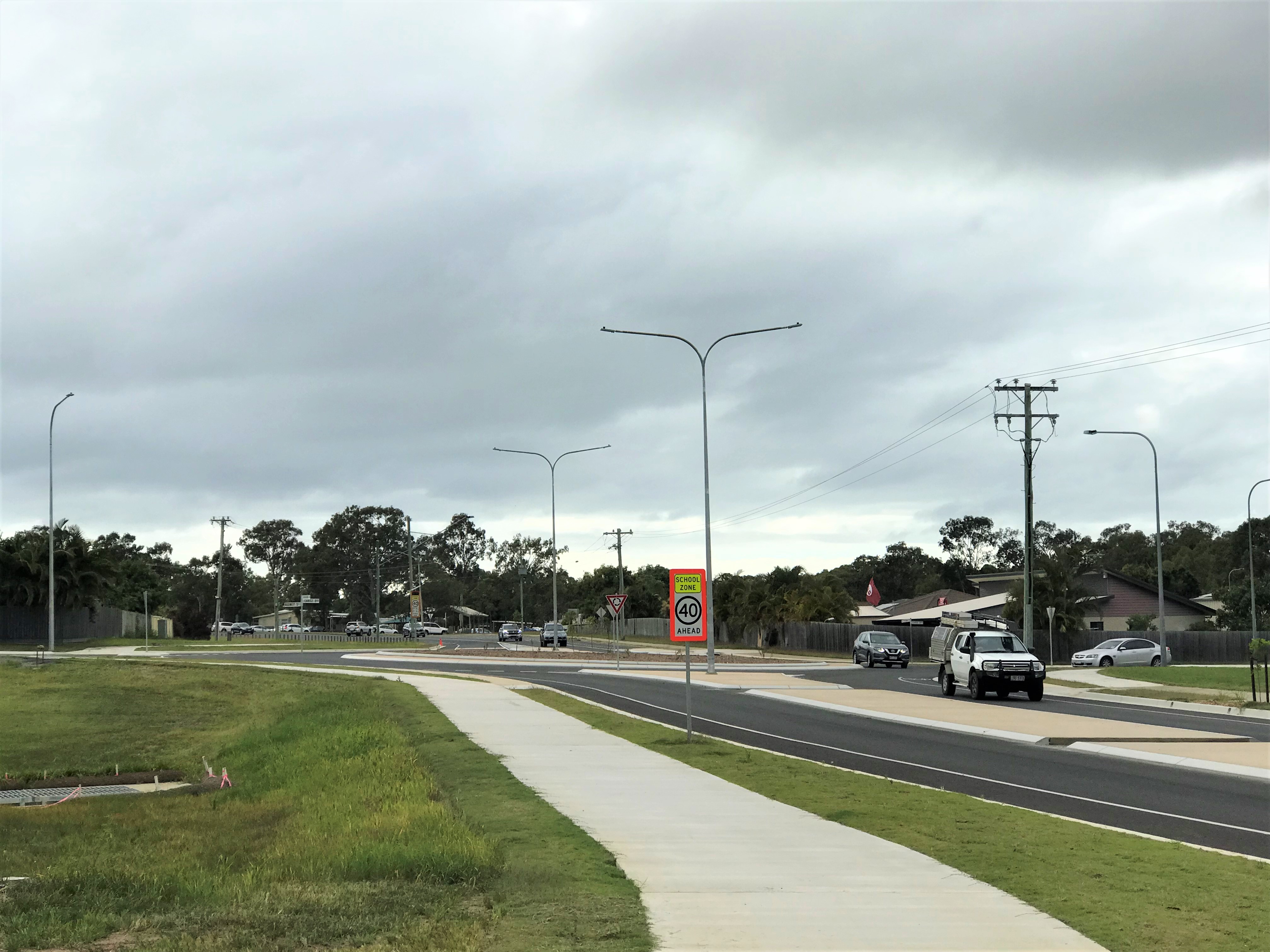 Carige Boulevard Roundabout with cars and a school zone sign showing a 40 km/h speed limit, alongside a sidewalk, trees, and houses under an overcast sky.