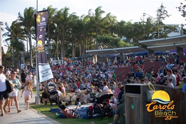 Crowd gathered at Yeppoon Main Beach for the 20th Carols by the Beach event, with palm trees and banners in the background.