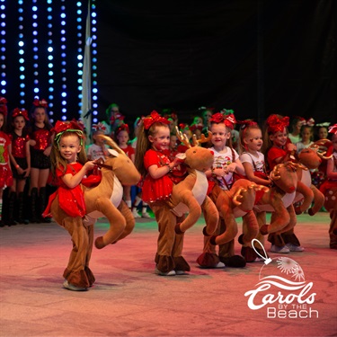 Children in red costumes with reindeer props performing on stage at a Carols by the Beach event.