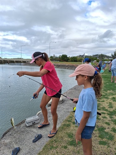 Two people fishing by the water; one holds a rod with a small fish. Others and fishing gear visible nearby under a cloudy sky.