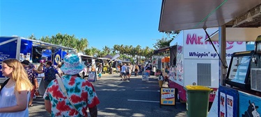 People walking through a sunny outdoor market with food trucks, including Yeppoon Lions Club and Mr. Whippy, and a sign reading “Cold drinks here.”