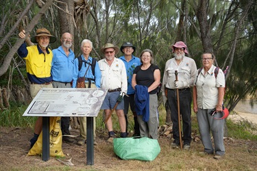 Eight people in outdoor clothing posing behind a sign titled “Beach scrub – an endangered community,” in a wooded area with tools and bags nearby.