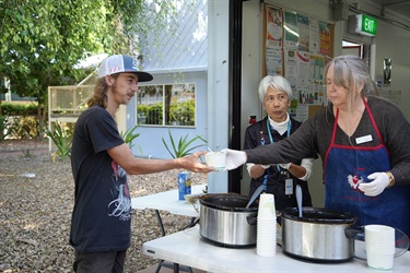 Person receiving food from two volunteers at a community service table with large pots, cups, and supplies, set outdoors near a building with posters.