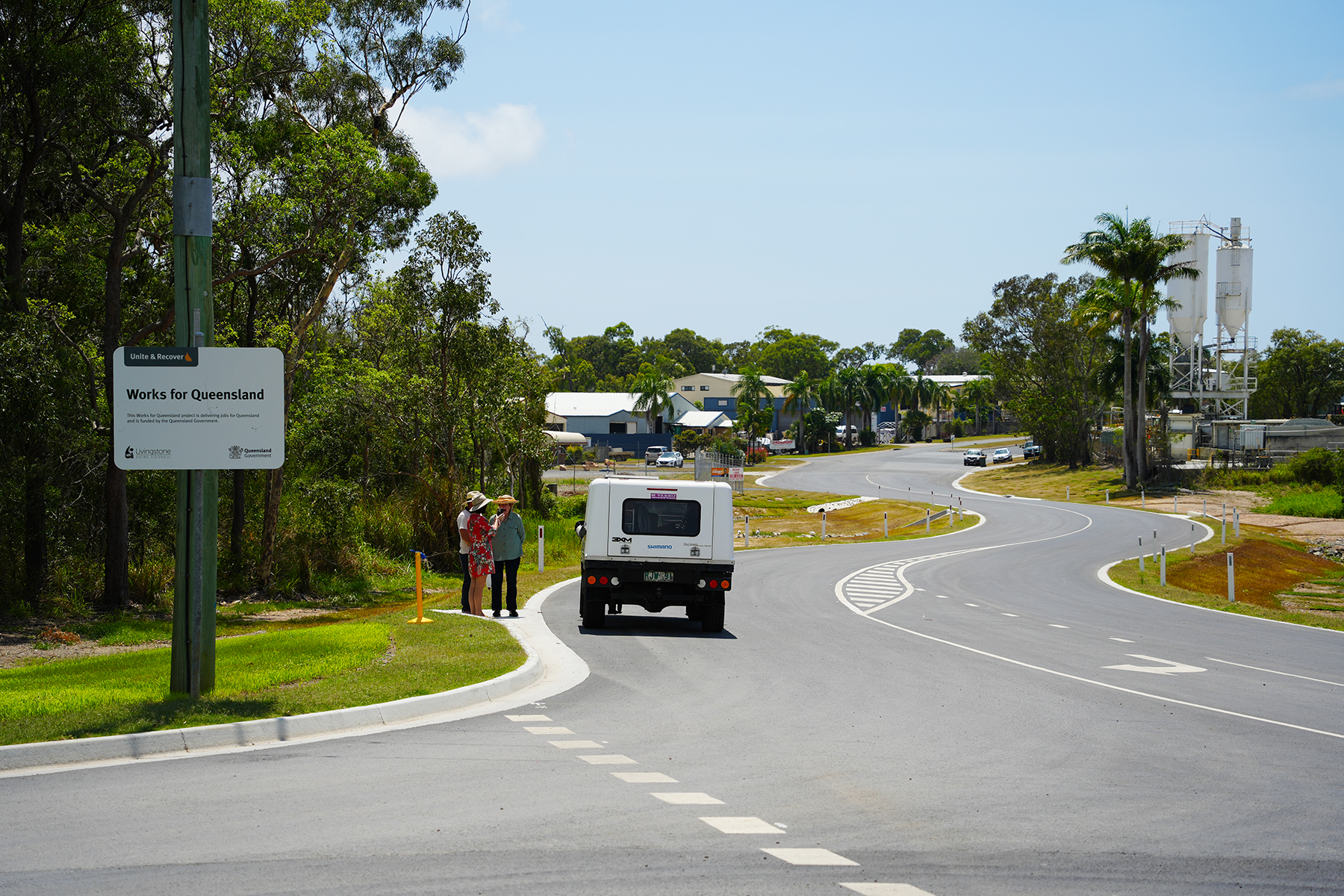 Three people standing near a “Works for Queensland” sign beside a newly constructed road, with a white vehicle and buildings in the background at Jabiru Drive Extension