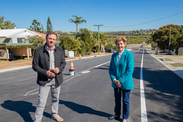 Two people standing on a newly paved residential road with traffic cones and houses on both sides under a clear blue sky.