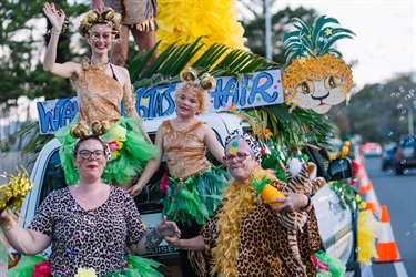 People in colourful costumes with leaf skirts and animal prints standing on a decorated vehicle with trees and traffic cones in the background.
