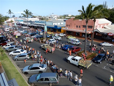Street parade with decorated vehicles and people walking, watched by crowds along a road lined with palm trees, parked cars, and an IGA store.