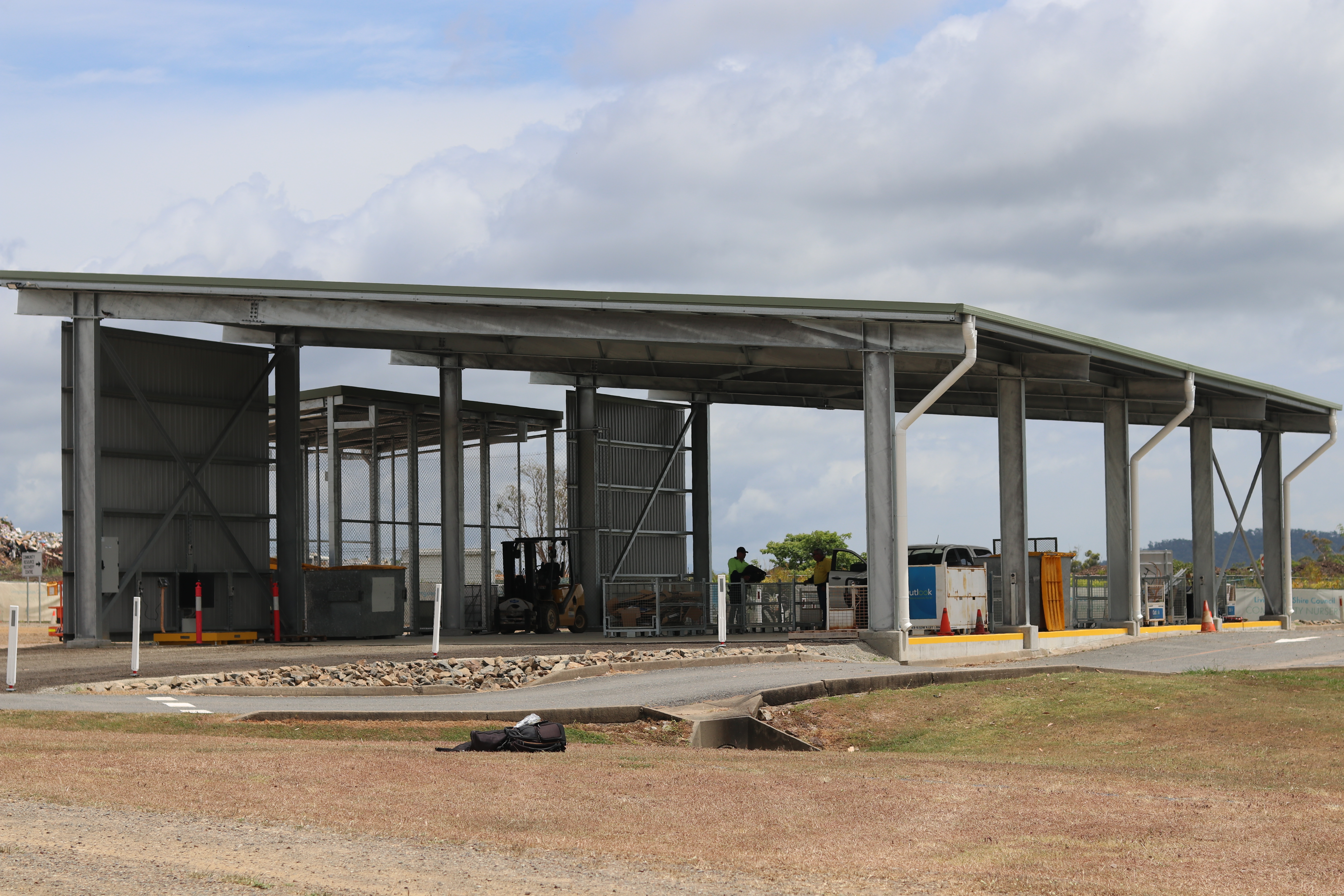 Two workers in high-visibility clothing inside an open-sided industrial structure with a forklift and containers, near a drainage ditch with rocks and grass.