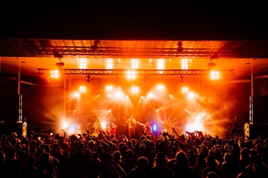 Band performing on a brightly lit stage with orange and yellow lights, as a large crowd raises their hands at a live concert.