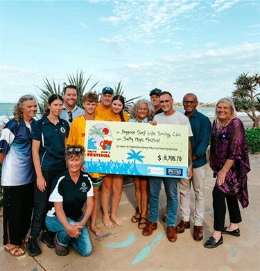 Group of people outdoors holding a $6,795.70 cheque from the Salty Hops Festival to Yeppoon Surf Life Saving Club, with palm trees and a blue sky in the background.