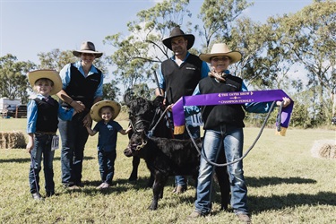 Group of people in western-style clothing standing on grass with a black calf, holding a ribbon that reads “The Caves Show – Champion Female.”