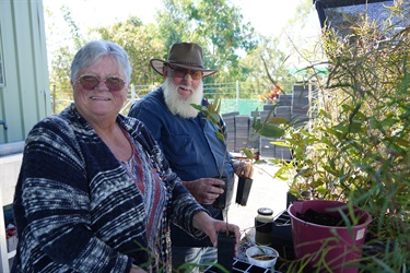 Two people outdoors holding potted plants near a table with gardening supplies, including a pink bucket, containers, and greenery in the background.