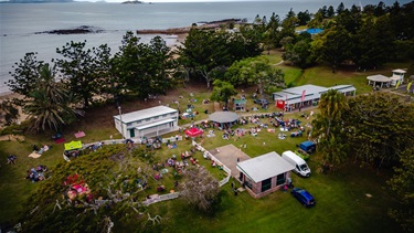 Aerial view of a park near the water with people gathered on the grass, some near tents and small buildings, suggesting a community event.