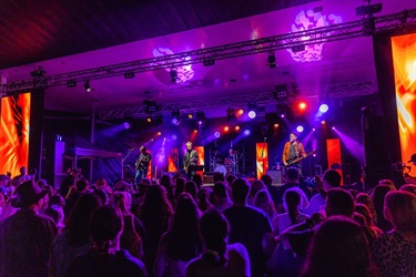 Band performing on a stage lit with purple and orange lights, with LED screens and a large crowd watching at a live concert.