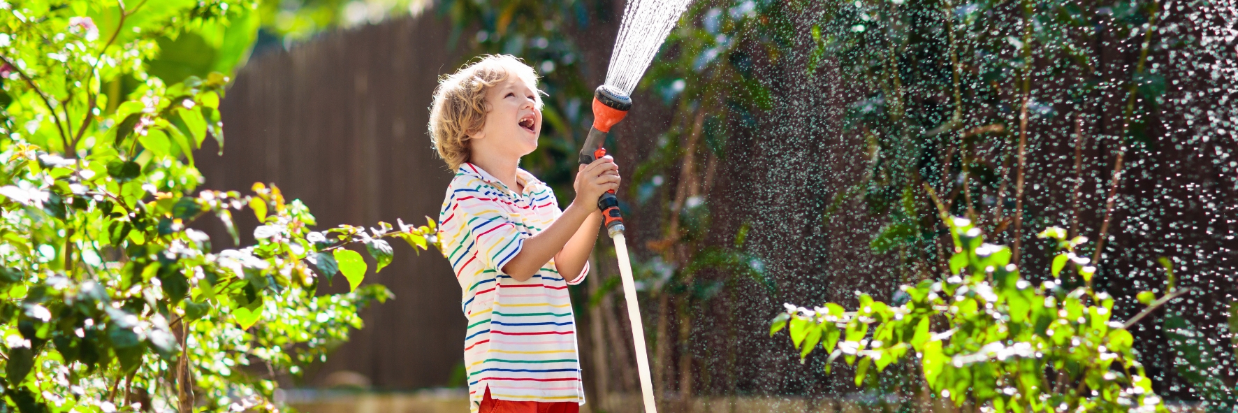 Child watering garden plants with a hose, surrounded by greenery and flowers.