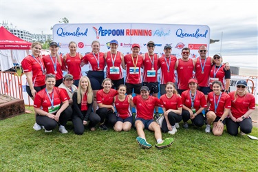 Group of people in red shirts and medals posing in front of a “Queensland Yeppoon Running Festival” backdrop near the beach.