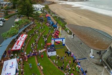 People gathered on a grassy area near the beach for an outdoor event with tents, banners, and a large curved-roof structure in view.