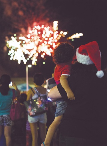 Person in a Santa hat holding a child while watching colourful fireworks at night, with others nearby enjoying the display.