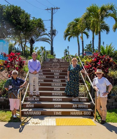 Four people standing beside outdoor steps featuring multilingual words related to nature, such as sun, moon, and rain.