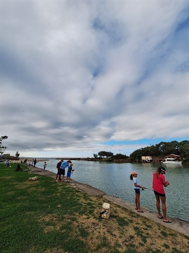 People fishing along a concrete path beside a river or canal, with boats, trees, and a small building in the background under a partly cloudy sky.