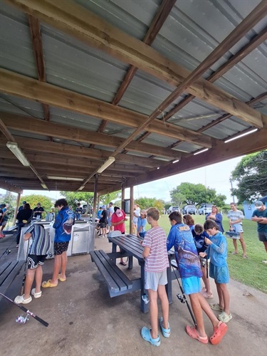 Group of adults and children under a covered outdoor area with picnic tables, holding fishing rods; trees and parked cars in the background.