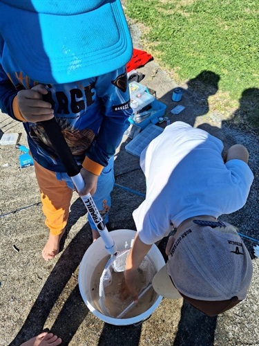 Two children outdoors with fishing gear; one holds a net, the other reaches into a white bucket of water.