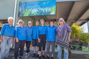 Group of six people in front of a building with a sign reading 'Home of Nursery Natives', surrounded by plants and gardening gear.