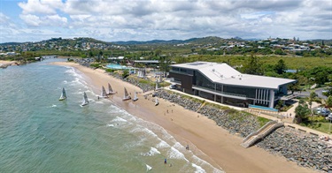 Sandy beach with small sailboats near the shore, people enjoying the water, and a modern building nearby, with a bridge and hillside houses in the background.